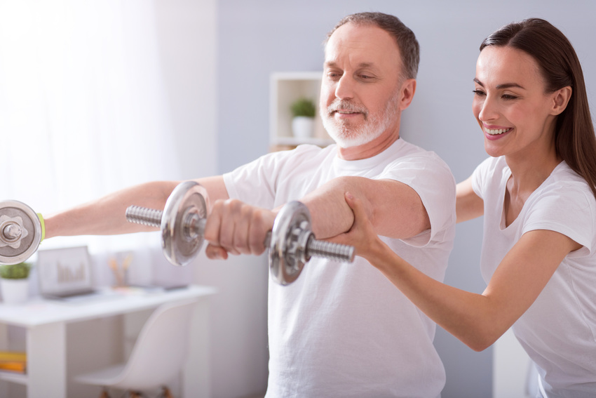 Using dumbbells. Cheerful male patient with dumbbells during physical therapy praxis and merry female physiotherapist helping him